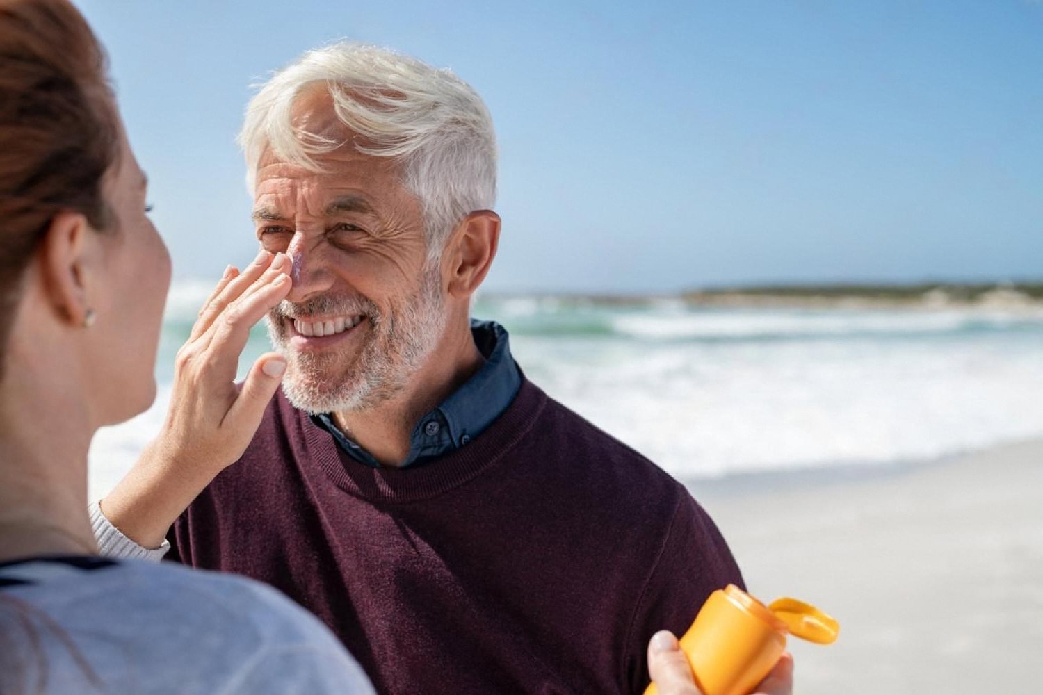 A woman applying sunscreen to a smiling man's nose on a sunny Dubai beach, showing daily sun protection for new UAE residents